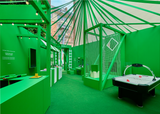 lifestyle: AVOLT pop-up showroom with green display booths, product counters, netted exhibit and air hockey table